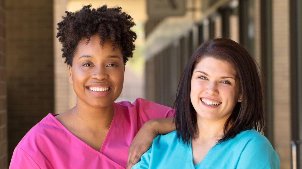 Two smiling nurses, one wearing pink scrubs, and one wearing blue scrubs