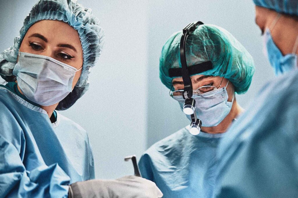 Three people in medical PPE stand in a circle in a surgery room.
